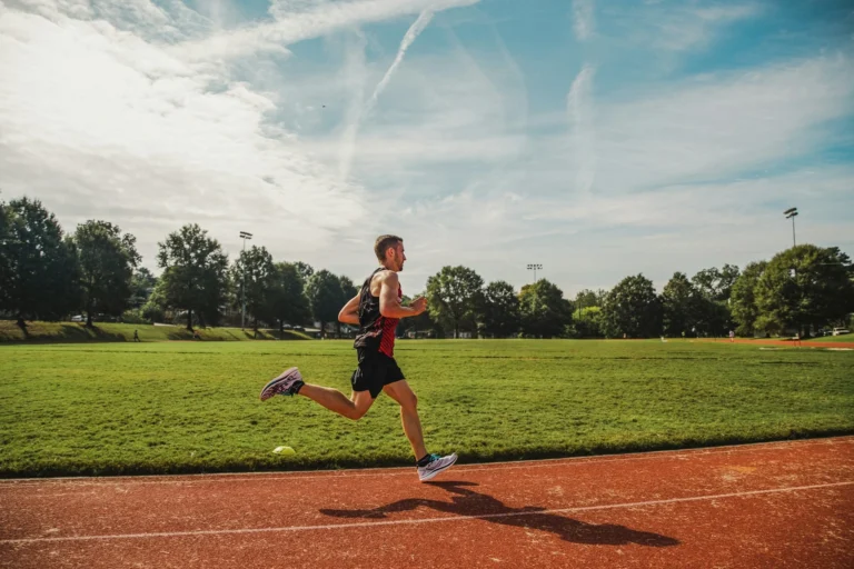 A male runner in athletic gear sprinting on a red outdoor track under a bright, sunny sky with scattered clouds.