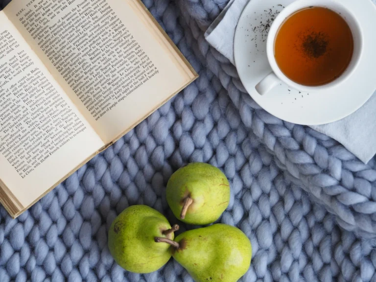 A flat-lay view of an open book, a cup of amber tea on a saucer, and three green pears resting on a thick, grey chunky-knit blanket.