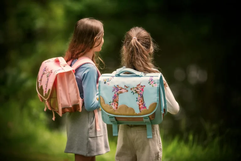 Two young girls are seen from behind, wearing decorative school backpacks while outdoors with a blurred green background.